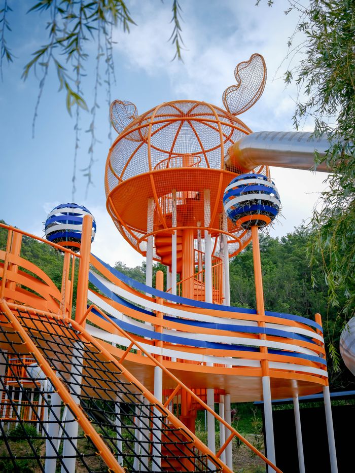 Colorful playground with climbing structures and slides, featuring orange and blue accents against a green backdrop.